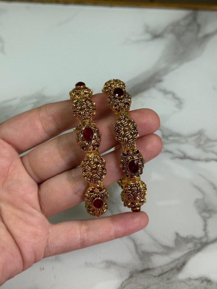 A pair of ornate antique gold bangles with large red oval gemstones and leaf-patterned goldwork, held in a hand over a marble background.