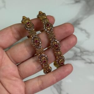 Close-up of intricately designed antique gold bangles held in hand, featuring floral filigree patterns and small pink gemstone embellishments.