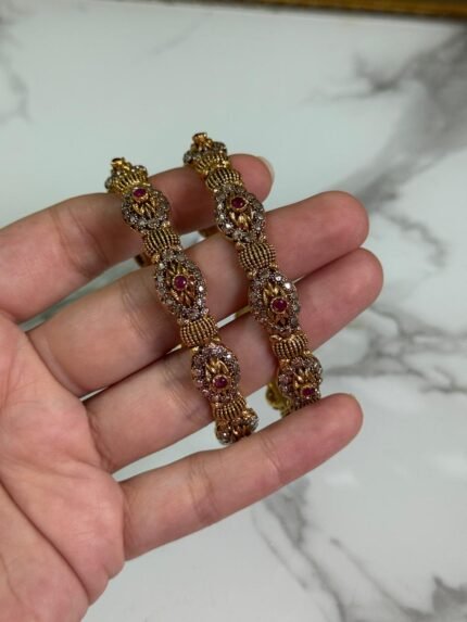 Close-up of intricately designed antique gold bangles held in hand, featuring floral filigree patterns and small pink gemstone embellishments.