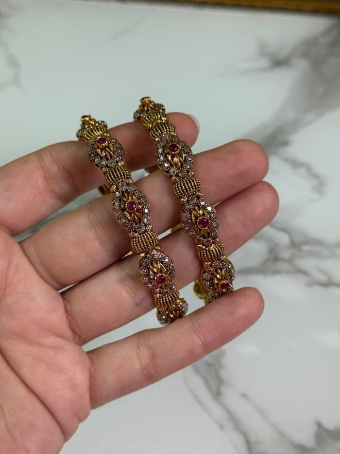 Close-up of intricately designed antique gold bangles held in hand, featuring floral filigree patterns and small pink gemstone embellishments.