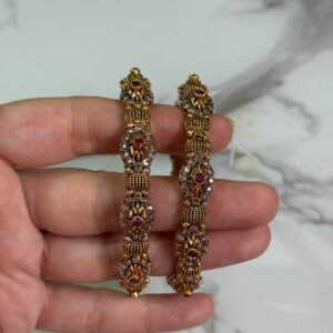Close-up of intricately designed antique gold bangles held in hand, featuring floral filigree patterns and small pink gemstone embellishments.