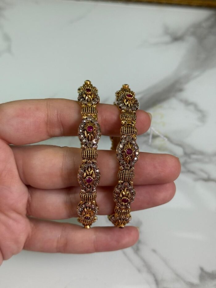 Close-up of intricately designed antique gold bangles held in hand, featuring floral filigree patterns and small pink gemstone embellishments.