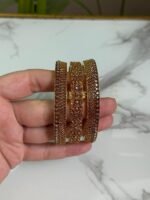 A hand holding a set of three gold bangles, each encrusted with detailed crystal patterns in symmetrical and wave designs, displayed against a marble background.