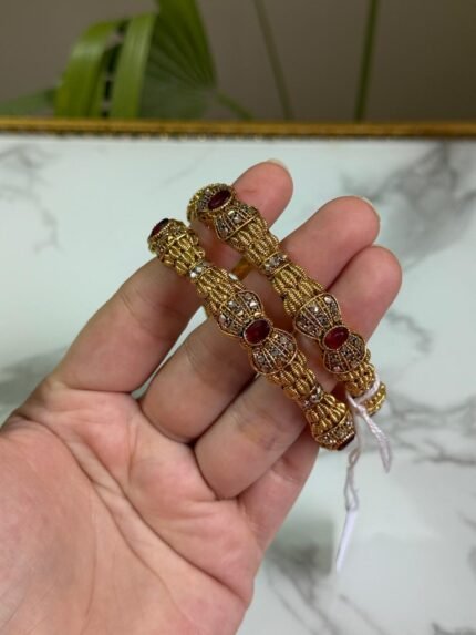 Hand holding a pair of traditional gold bangles with detailed textured design and oval red gemstones, placed against a marble background.