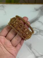 A close-up image of a hand holding three intricately designed gold bangles, featuring detailed floral and filigree patterns, against a marble background.