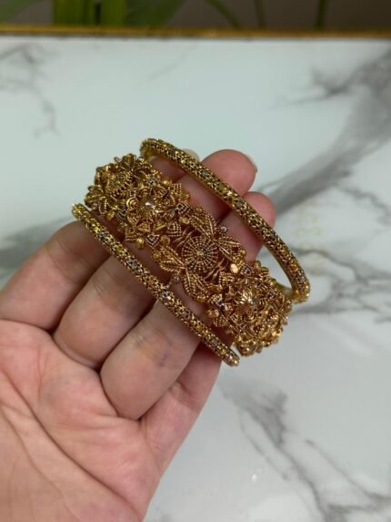A close-up image of a hand holding three intricately designed gold bangles, featuring detailed floral and filigree patterns, against a marble background.