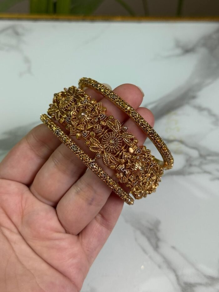 A close-up image of a hand holding three intricately designed gold bangles, featuring detailed floral and filigree patterns, against a marble background.