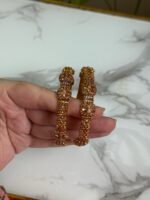 A pair of traditional gold bangles with rope-texture design and ornate bead detailing, held in hand over a marble surface.