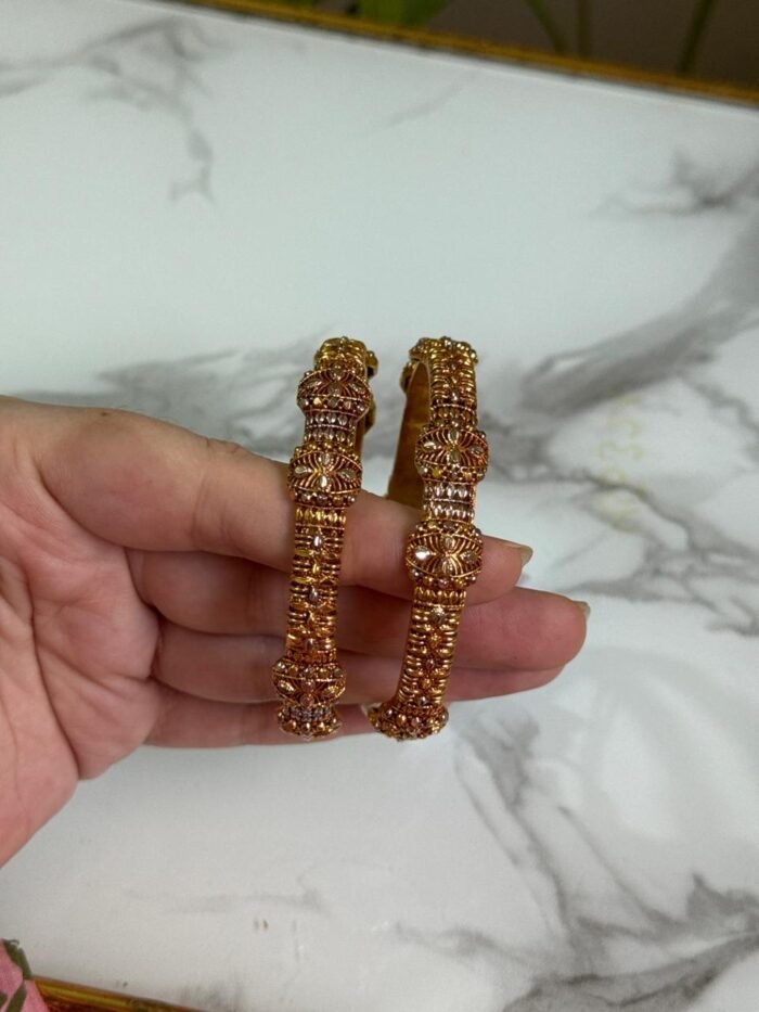 A pair of traditional gold bangles with rope-texture design and ornate bead detailing, held in hand over a marble surface.
