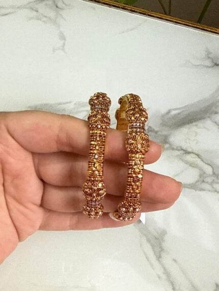 A pair of traditional gold bangles with rope-texture design and ornate bead detailing, held in hand over a marble surface.