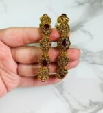 A pair of ornate antique gold bangles with large red oval gemstones and leaf-patterned goldwork, held in a hand over a marble background.