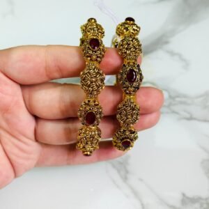 A pair of ornate antique gold bangles with large red oval gemstones and leaf-patterned goldwork, held in a hand over a marble background.