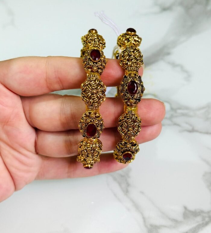A pair of ornate antique gold bangles with large red oval gemstones and leaf-patterned goldwork, held in a hand over a marble background.