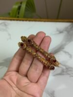 A hand holding a pair of antique-finish gold bangles with intricate floral carvings and red oval center stones, displayed against a marble background.