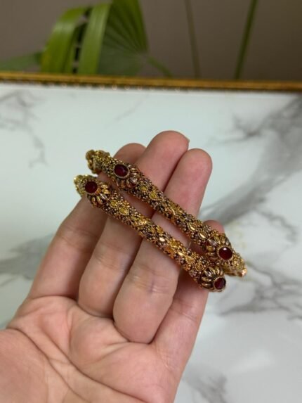 A hand holding a pair of antique-finish gold bangles with intricate floral carvings and red oval center stones, displayed against a marble background.