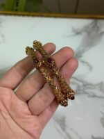 A hand holding a pair of antique-finish gold bangles with intricate floral carvings and red oval center stones, displayed against a marble background.