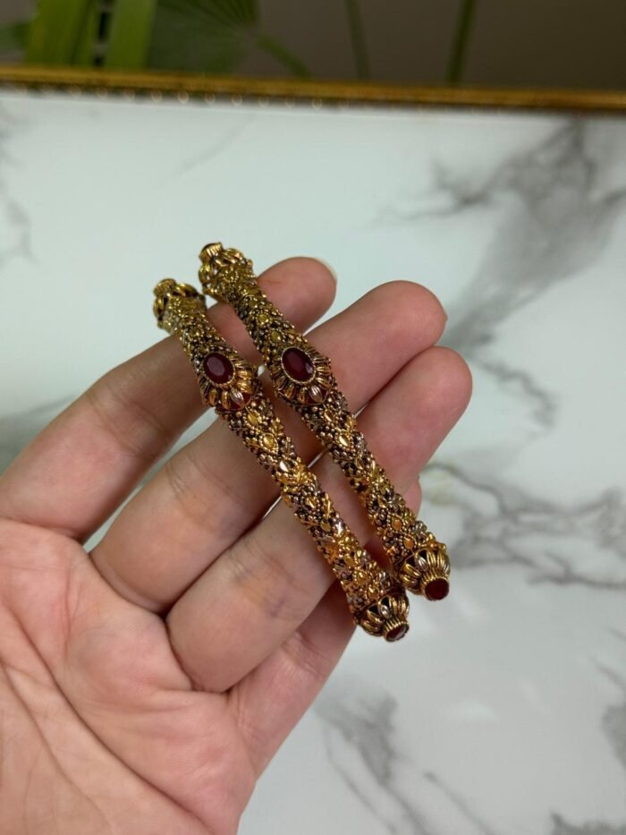 A hand holding a pair of antique-finish gold bangles with intricate floral carvings and red oval center stones, displayed against a marble background.