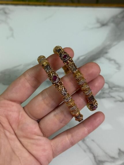 A close-up of a hand holding a pair of gold-plated ethnic bangles with intricate filigree patterns and ruby red stone embellishments, against a marble background.