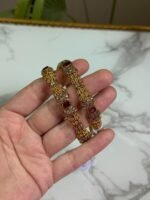 Hand holding a pair of traditional gold bangles with detailed textured design and oval red gemstones, placed against a marble background.