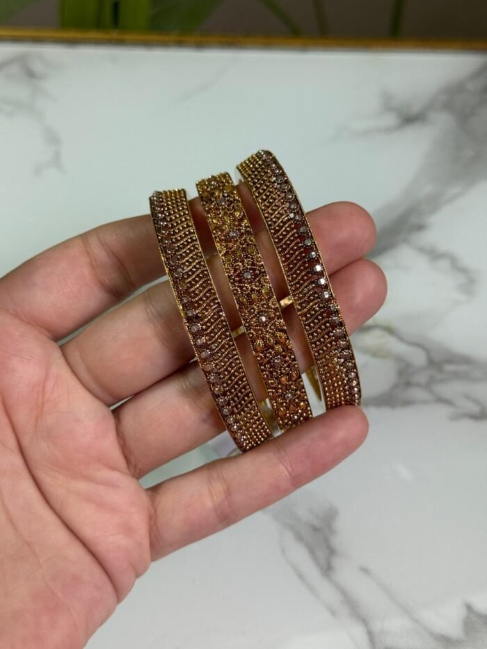 A hand holding a set of three gold bangles, each encrusted with detailed crystal patterns in symmetrical and wave designs, displayed against a marble background.