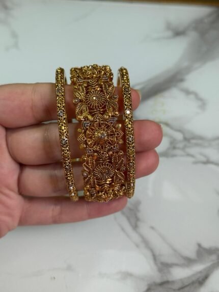 A close-up image of a hand holding three intricately designed gold bangles, featuring detailed floral and filigree patterns, against a marble background.