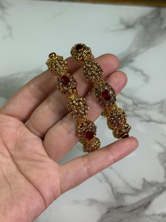 A pair of ornate antique gold bangles with large red oval gemstones and leaf-patterned goldwork, held in a hand over a marble background.