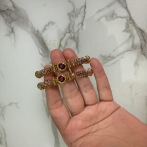 A pair of antique gold bangles with intricate textured design and round pink gemstone centers, held in hand against a marble backdrop.