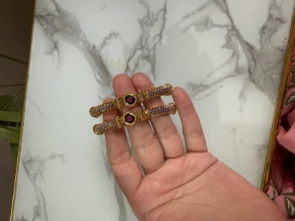 A pair of antique gold bangles with intricate textured design and round pink gemstone centers, held in hand against a marble backdrop.