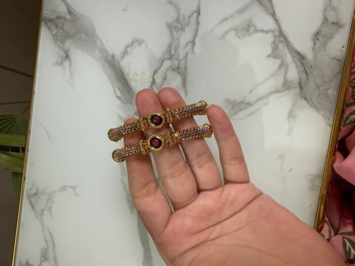 A pair of antique gold bangles with intricate textured design and round pink gemstone centers, held in hand against a marble backdrop.