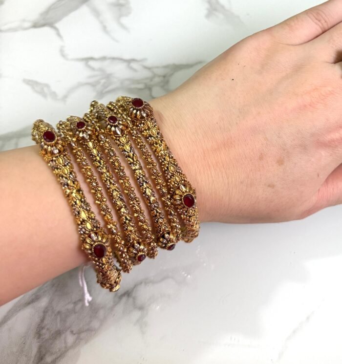 A hand adorned with a set of six antique gold bangles, each intricately designed with ornate patterns and embedded with oval-shaped deep red stones