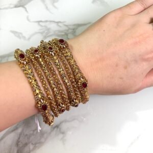 A hand adorned with a set of six antique gold bangles, each intricately designed with ornate patterns and embedded with oval-shaped deep red stones