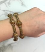 Wrist adorned with two antique-style gold bangles featuring floral motifs and detailed filigree work, set against a marble background.