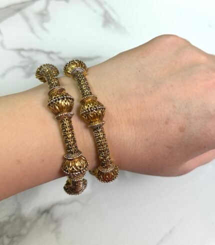 Wrist adorned with two antique-style gold bangles featuring floral motifs and detailed filigree work, set against a marble background.