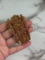 A close-up image of a hand holding three intricately designed gold bangles, featuring detailed floral and filigree patterns, against a marble background.