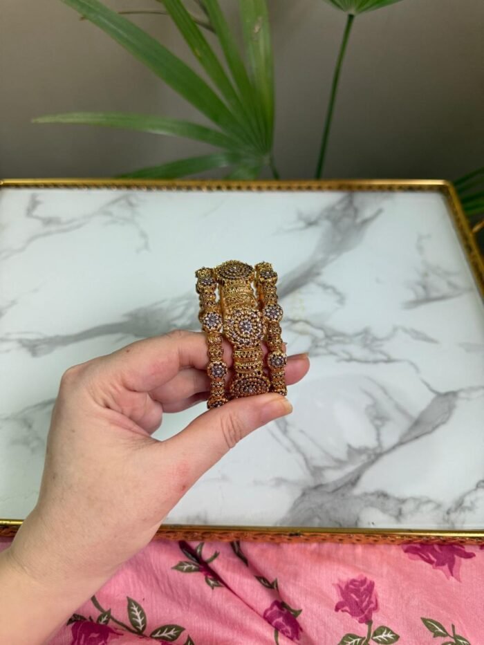 A pair of antique gold bangles featuring large round floral motifs encrusted with pink and white CZ stones, held in a hand over a marble background.
