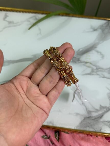 A hand holding two intricately crafted gold bangles featuring antique-style filigree work and floral motifs, with a marble surface in the background.