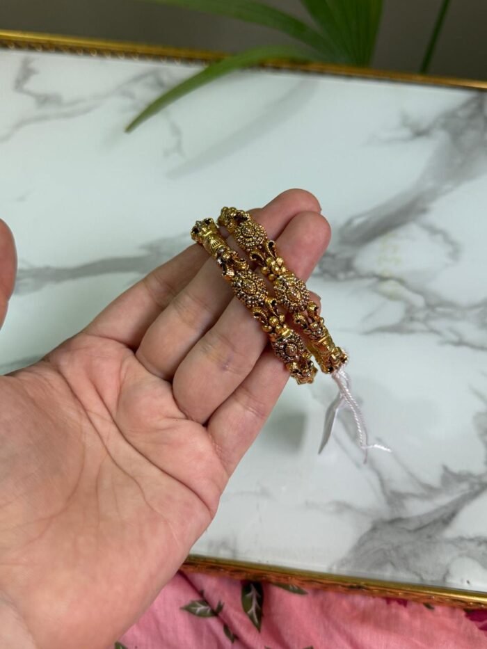 A hand holding two intricately crafted gold bangles featuring antique-style filigree work and floral motifs, with a marble surface in the background.