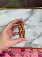 A close-up of a hand holding two antique-style gold bangles, featuring delicate maroon stone embellishments and detailed filigree patterns, displayed against a white marble background and pink floral fabric. Ideal for traditional or festive occasions.