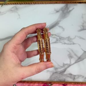 A close-up of a hand holding two antique-style gold bangles, featuring delicate maroon stone embellishments and detailed filigree patterns, displayed against a white marble background and pink floral fabric. Ideal for traditional or festive occasions.