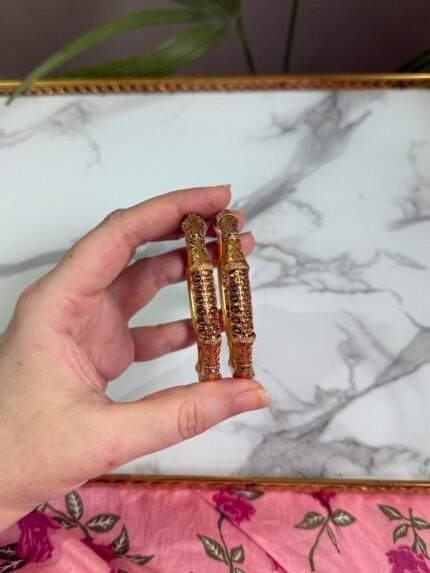 A close-up of a hand holding two antique-style gold bangles, featuring delicate maroon stone embellishments and detailed filigree patterns, displayed against a white marble background and pink floral fabric. Ideal for traditional or festive occasions.