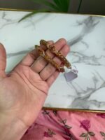 Hand holding a pair of antique-style gold bangles featuring ruby-colored center stones and intricate beaded detailing, placed over a marble surface with a pink floral fabric in the background.