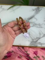 Hand holding a pair of antique-style gold bangles featuring ruby-colored center stones and intricate beaded detailing, placed over a marble surface with a pink floral fabric in the background.
