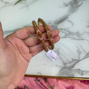 Hand holding a pair of antique-style gold bangles featuring ruby-colored center stones and intricate beaded detailing, placed over a marble surface with a pink floral fabric in the background.