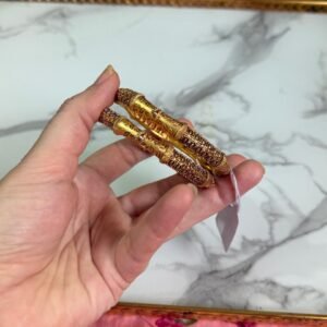 A close-up of a hand holding two antique-style gold bangles, featuring delicate maroon stone embellishments and detailed filigree patterns, displayed against a white marble background and pink floral fabric. Ideal for traditional or festive occasions.