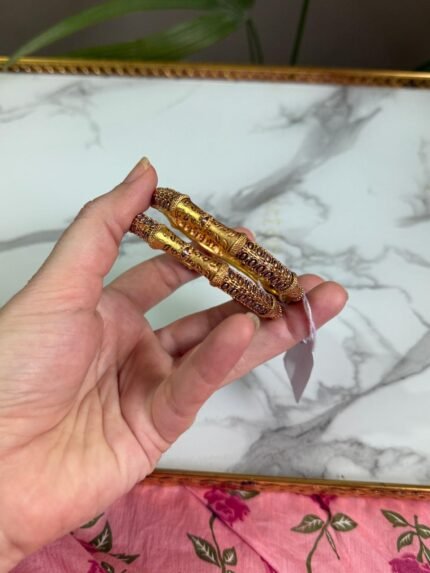 A close-up of a hand holding two antique-style gold bangles, featuring delicate maroon stone embellishments and detailed filigree patterns, displayed against a white marble background and pink floral fabric. Ideal for traditional or festive occasions.