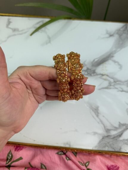 Hand holding a pair of traditional gold-tone bangles with floral cluster motifs and intricate detailing, placed over a marble surface.