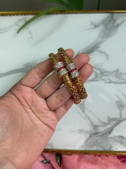 A hand holding two gold-toned bangles with intricate floral patterns and sparkling stone-studded accents in silver and ruby hues, displayed against a marble background.
