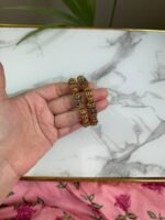 A hand holding a pair of antique gold-toned bangles with intricate floral filigree work and beaded textures, placed against a marble background with a touch of greenery.
