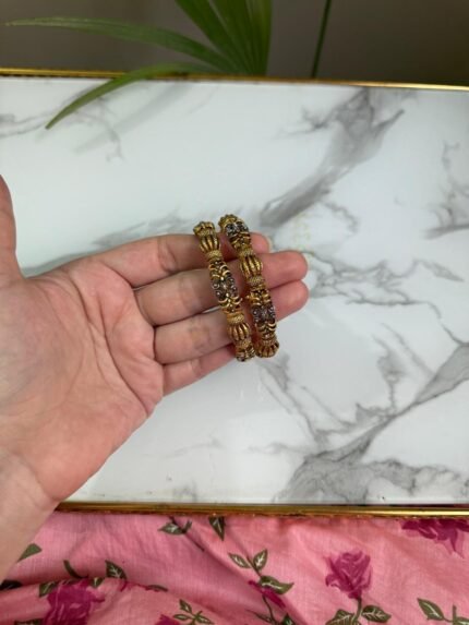 A hand holding a pair of antique gold-toned bangles with intricate floral filigree work and beaded textures, placed against a marble background with a touch of greenery.