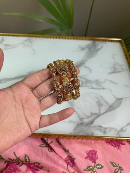 A pair of antique gold bangles featuring large round floral motifs encrusted with pink and white CZ stones, held in a hand over a marble background.
